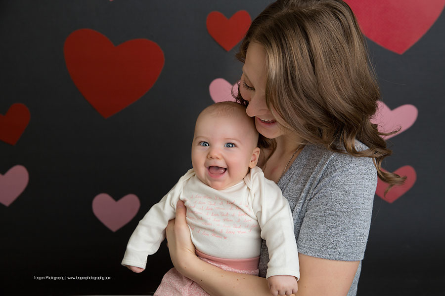 A blond haired mother snuggles with her baby in front of a grey wall covered in hearts