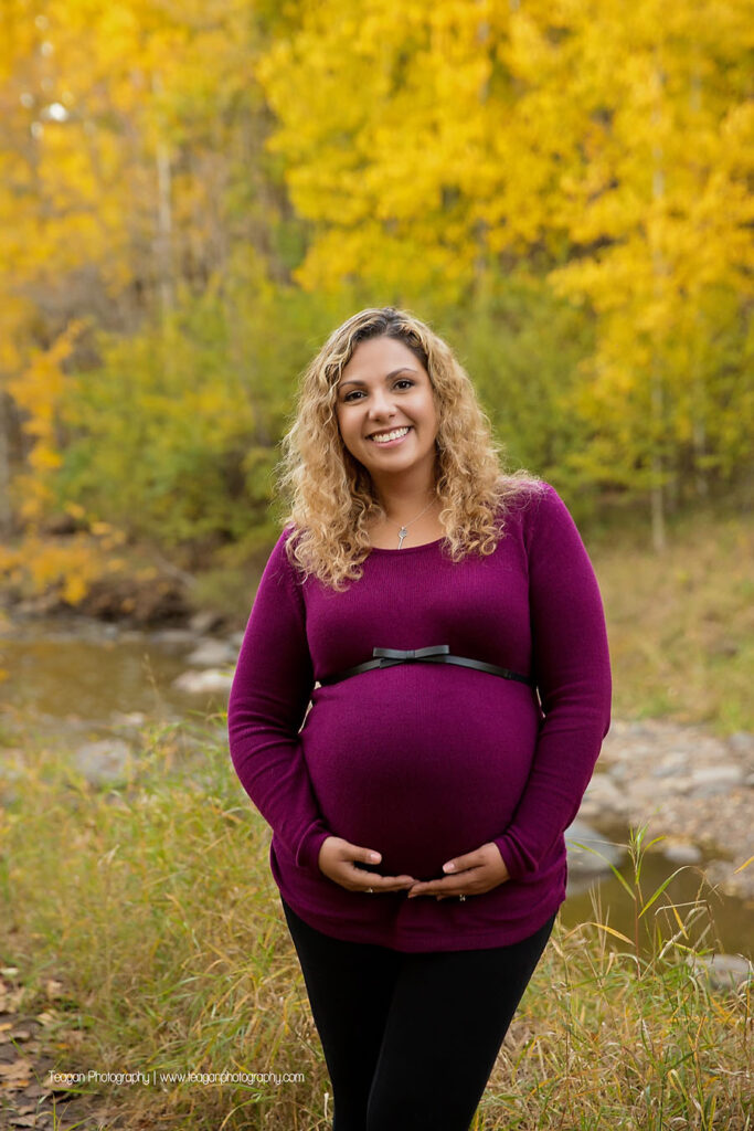 Close up of baby bump with a crisp running stream and colorful autumn leaves in the background in Edmonton