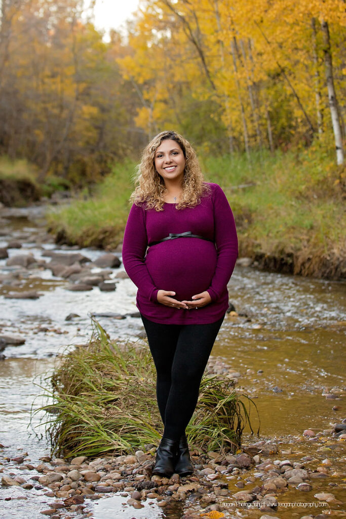 Pregnant mother walking along a rocky creek bed surrounded by golden autumn leaves in an Edmonton ravine